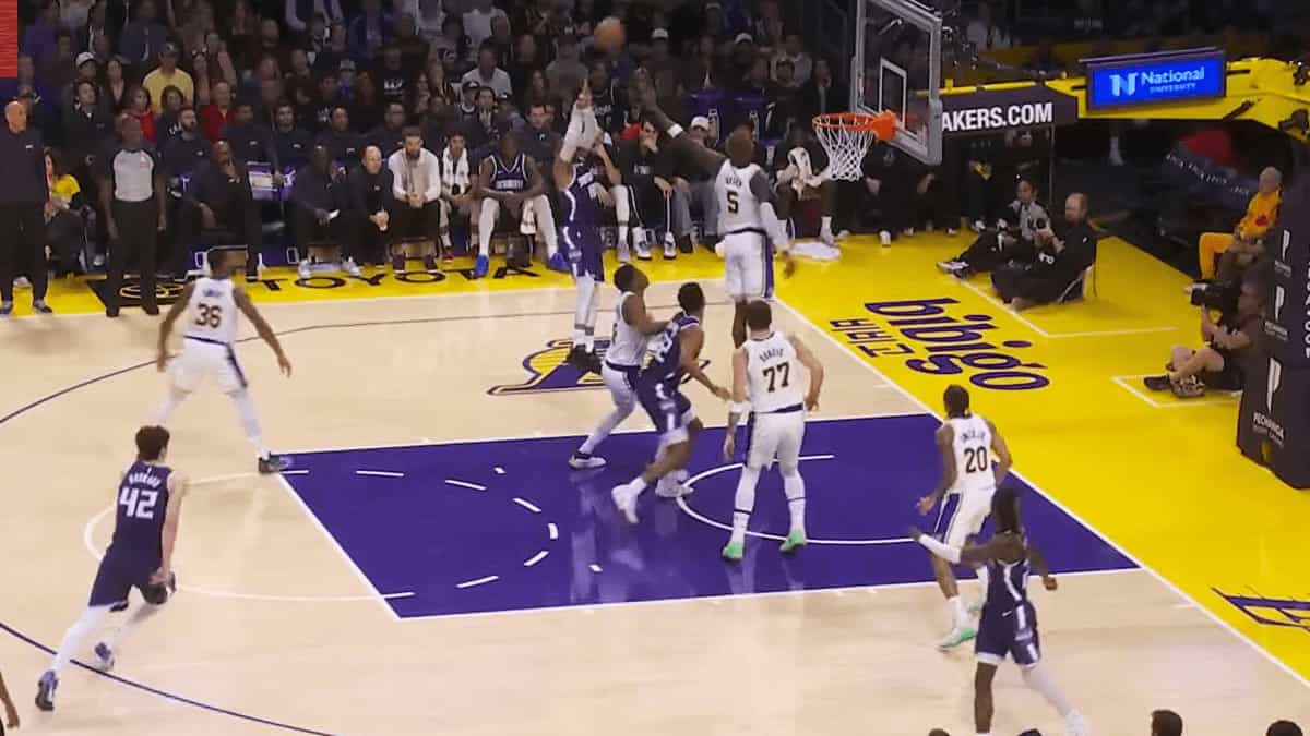 A Los Angeles Lakers defender contests a driving layup at the rim against a Sacramento Kings player during an NBA game, with teammates and crowd visible in the background.