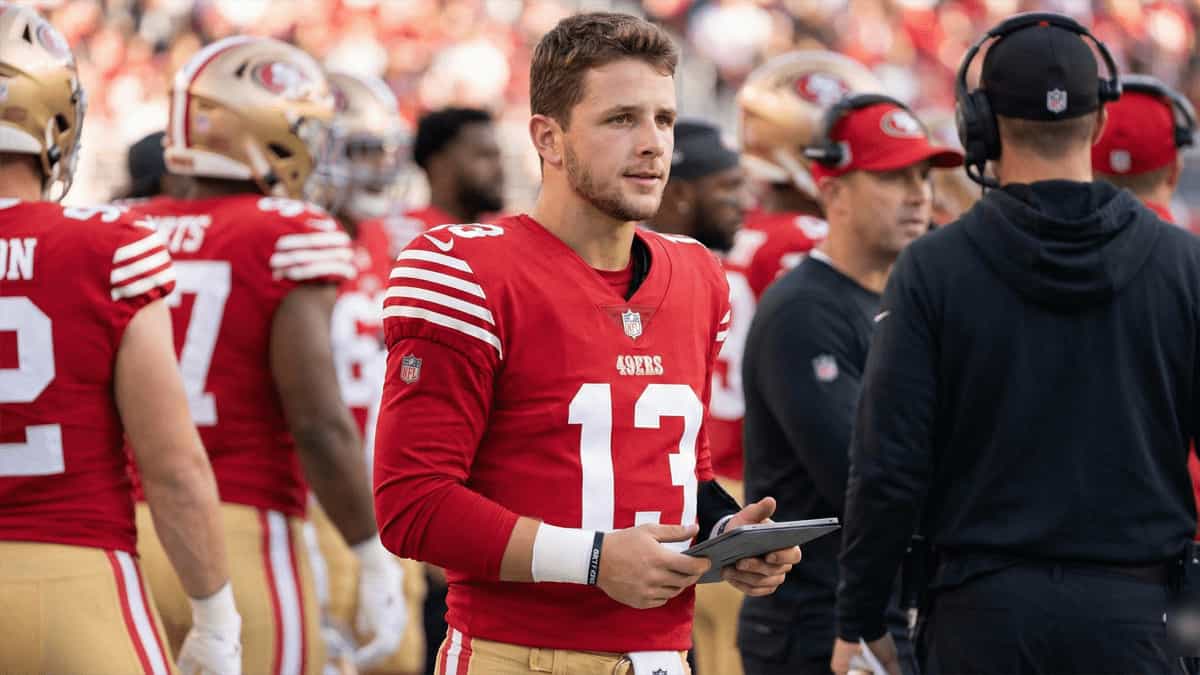 A quarterback wearing the number 13 jersey for the San Francisco 49ers stands on the sideline during an NFL game, holding a tablet while teammates and coaches gather in the background.